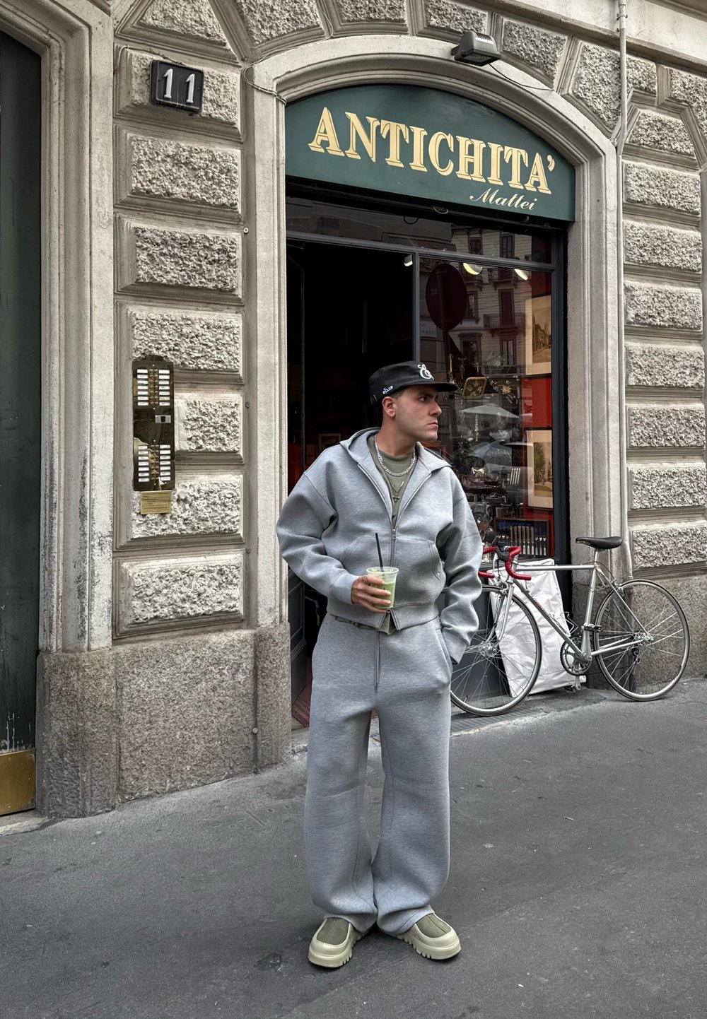 Szary, oversize'owy sweatshirt i dopasowane spodnie z miękkiego materiału, z zamkiem i kieszeniami. Postać trzyma zielony napój i nosi ciemną czapkę.