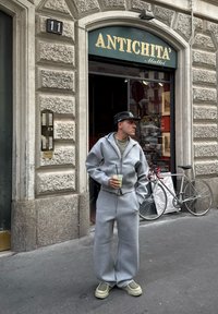 Szary, oversize'owy sweatshirt i dopasowane spodnie z miękkiego materiału, z zamkiem i kieszeniami. Postać trzyma zielony napój i nosi ciemną czapkę.