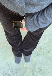 Černé manšestrové kalhoty, šedý pletený svetr a sluneční brýle se zlatými detaily držené v ruce. Šedé kotníkové boty s špičatou špičkou doplňují vzhled.