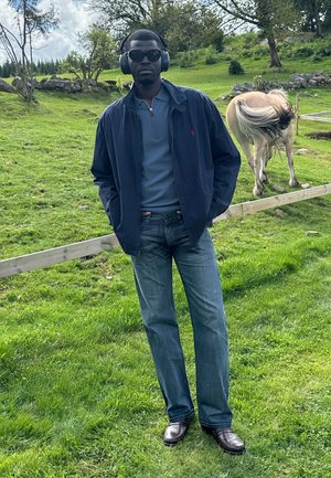 Hombre con una chaqueta navy sobre un polo azul claro, usando gafas de sol y auriculares. De pie sobre la hierba, con un caballo beige de fondo.