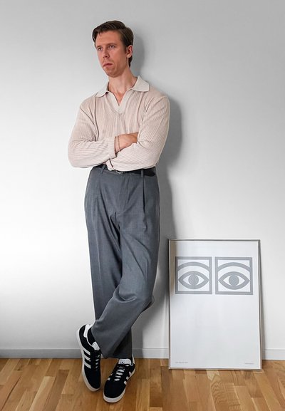 Jersey de punto texturizado en beige claro, con un cuello blanco, combinado con pantalones grises con pliegues y zapatillas negras. Obra de arte enmarcada en el suelo.