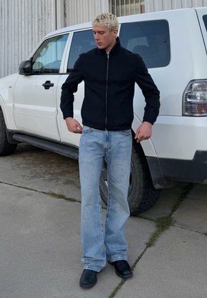 Chaqueta negra con cremallera, vaqueros acampanados de color azul claro, zapatos negros. De pie al aire libre cerca de un vehículo blanco, con asfalto texturizado y hierba.