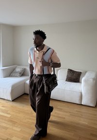 Homme portant un polo rayé et un pantalon marron, tenant un sac en cuir noir, debout sur un sol en bois dans un salon minimaliste.