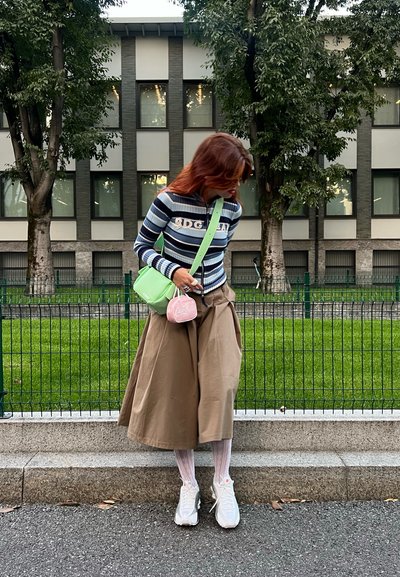 Camisa de manga larga a rayas, falda midi beige, zapatillas blancas, bolso cruzado verde, monedero rosa. Al aire libre con un fondo de vegetación y edificio.