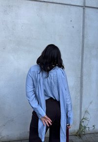 Camisa azul claro de manga longa com detalhe franzido nas costas, combinada com calças escuras; em pé contra uma parede de betão; mãos posicionadas atrás das costas.