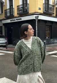 A woman with short dark hair, wearing a green knitted cardigan and a white lace skirt, stands at a street corner near a yellow building with balconies.