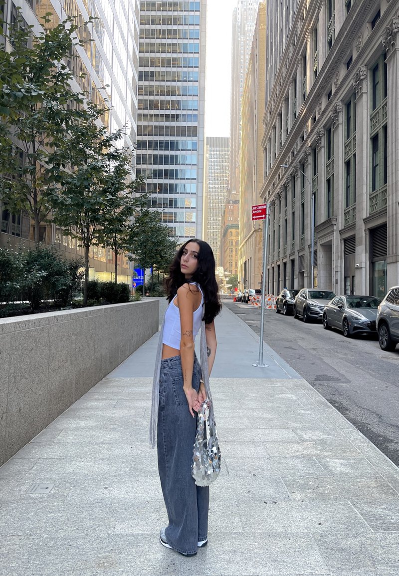 Γκρίζα παντελόνια σε φαρδιά γραμμή, λευκή κοντομάνικη μπλούζα και τσάντα με ασημένια παγιέτα, μπροστά σε μια υφασμάτινη αστική τοπίο με δέντρα κατά μήκος του πεζοδρομίου.