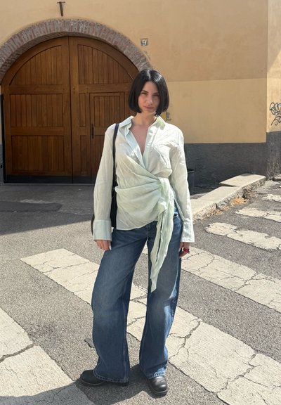 Mujer con cabello corto bob negro, que lleva una blusa cruzada verde claro, vaqueros de pierna ancha y zapatos negros, de pie en un paso de peatones frente a una puerta arqueada de madera.