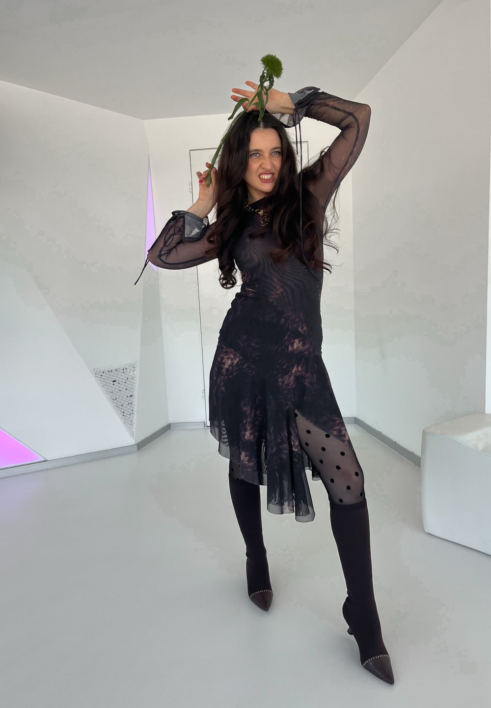 Schwarzes, transparentes Kleid mit Polka-Dots, hohem Schlitz und langen Ärmeln. Kombiniert mit knielangen, enganliegenden schwarzen Socken und spitzen Absätzen. Grüner Blumenakzent.