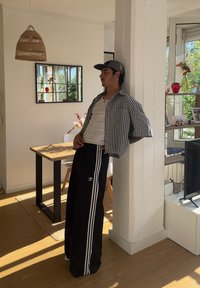 Conjunto casual com uma camisa de manga curta xadrez, uma t-shirt branca em baixo e calças de treino pretas com três riscas brancas e logotipo. Ambiente interior bem iluminado.