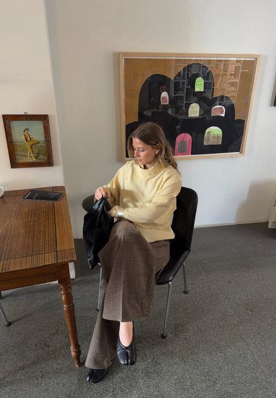 Mujer con suéter beige y pantalones marrones sentada en una silla negra junto a una mesa de madera, sosteniendo una bolsa negra, con pinturas abstractas y clásicas en la pared detrás.