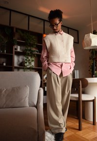 Gilet en tricot beige sur une chemise boutonnée rose clair, associé à un pantalon large beige et des chaussures noires. Textures douces et design superposé.