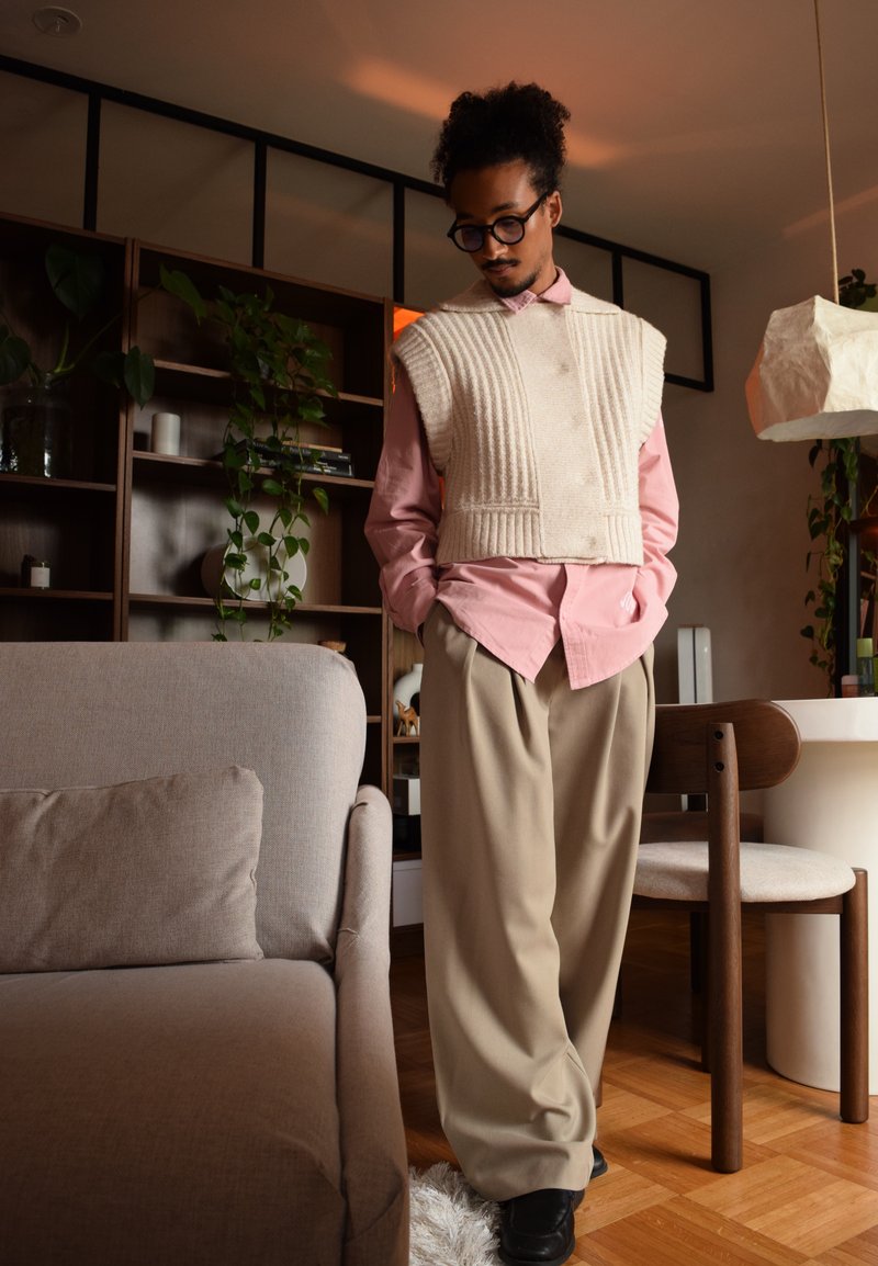 Gilet en tricot beige sur une chemise boutonnée rose clair, associé à un pantalon large beige et des chaussures noires. Textures douces et design superposé.
