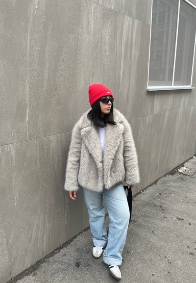 Mujer con un gorro rojo, gafas de sol negras, abrigo de piel sintética beige, jeans azul claro y zapatillas blancas, de pie junto a una pared texturizada gris.