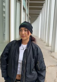 Νέος που φορά μαύρο σκουφάκι και μπουφάν στέκεται σε έναν σύγχρονο διάδρομο με λευκούς κίονες και μεγάλα παράθυρα από τη μία πλευρά.