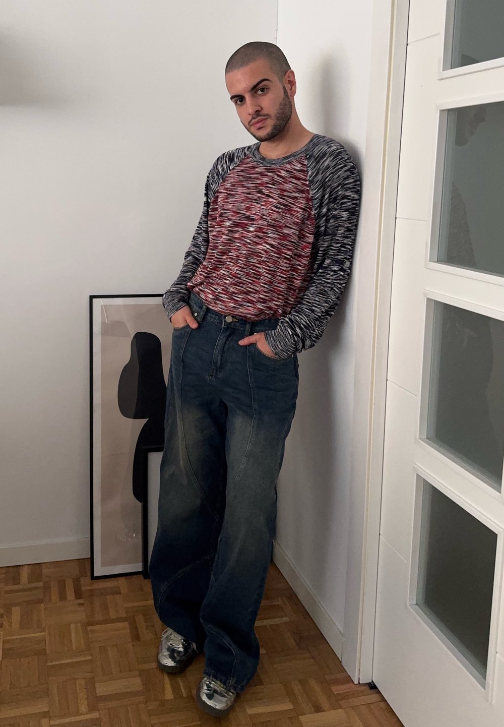Szürke, textúrázott, hosszú ujjú ing piros kiegészítőkkel, széles szárú kék farmernadrággal és fémes ezüst cipővel párosítva. Fal mellett áll.
