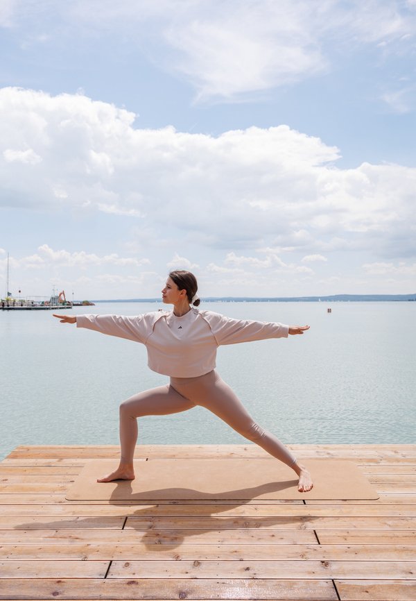 Sudadera corta de color rosa claro y leggins a juego, llevados por una persona en una postura de yoga sobre una esterilla marrón, con una plataforma de madera debajo y cerca del agua.