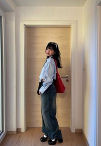 Una giovane donna con lunghi capelli neri si trova all'interno indossando una maglietta corta blu chiaro, jeans larghi, scarpe con tacco nero, calzini bianchi e una borsa rossa a tracolla.
