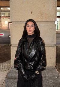 Zwarte leren jas met een hoge kraag en volumineuze mouwen, gecombineerd met een nauwsluitende zwarte top en een lange vloeiende rok. Handschoenen maken de look compleet.