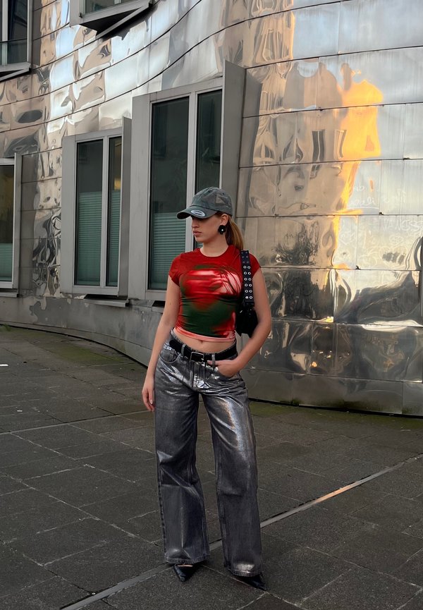 Rotes und grünes enges T-Shirt mit abstrakten Mustern, metallische weit geschnittene Jeans, schwarzer Gürtel und Camouflage-Cap, vor einer reflektierenden Gebäudefassade.