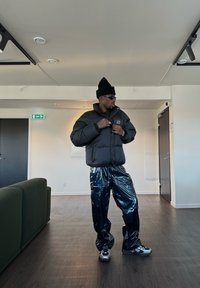 Svart dunjakke med høy krage, skinnende svarte vinylbukser og grå sneakers. Accessorisert med en svart lue og solbriller.