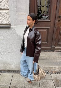 Veste en faux cuir marron avec un col haut, top gris clair et jeans larges bleu clair. Sac à main en crochet et chaussures beiges.