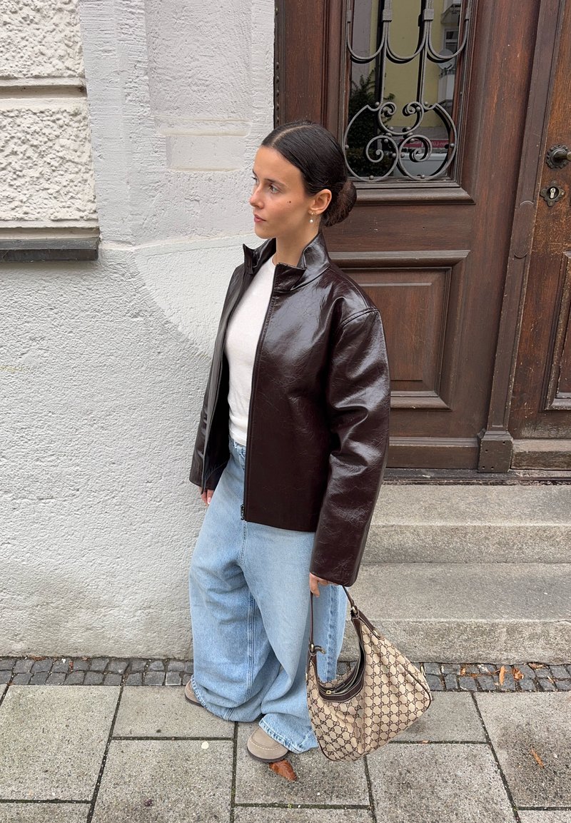 Brown faux leather jacket with a high collar, light gray top, and light blue wide-leg jeans. Crocheted handbag and beige shoes.