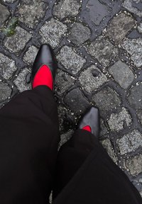 Chaussures noires à bout pointu avec un design ajouré, associées à des chaussettes rouges, sur un pavé mouillé. Pantalon sombre visible.