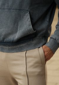 Mikina s kapucí v bledě modré látce s přední kapsou, doplněná béžovými kalhotami s bočními švy a hladkou texturou.