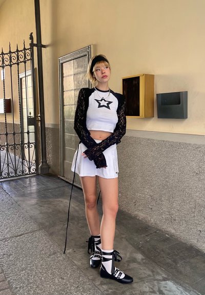 Las mangas largas de encaje negro enmarcan una blusa blanca corta con una estrella negra, combinada con una falda blanca y zapatos negros con correas y detalles.