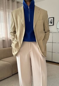 Blazer beige sur un col roulé côtelé bleu avec une fermeture éclair, associé à un pantalon large rose clair. Tissu lisse et design structuré.