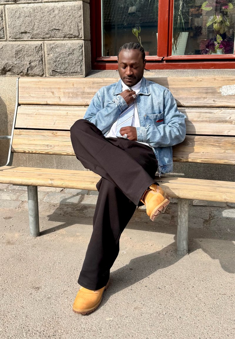 Homme aux cheveux tressés portant une veste en jean, une cravate, un pantalon noir et des bottes jaunes, assis les jambes croisées sur un banc en bois en extérieur.