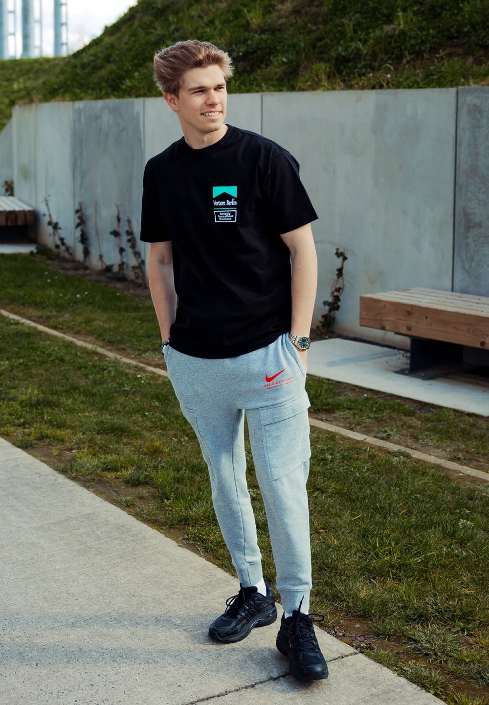Juoda marškinėliai su grafiniu logotipu ir tekstu, pilkos sportinės kelnės su raudonu Nike logotipu ir juodi sportbačiai. Stovi ant takelio šalia žolės.