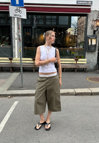 Una blusa blanca sin mangas, pantalones cortos cargo anchos en color verde oliva y chanclas negras. Los accesorios incluyen collares en capas y pulseras.