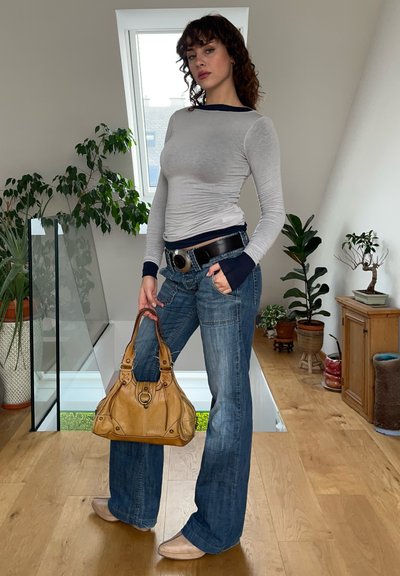 Camisa gris de manga larga con puños en azul marino, vaqueros acampanados azules, bolso color tostado con herrajes circulares y zapatos de color claro frente a un fondo neutro.