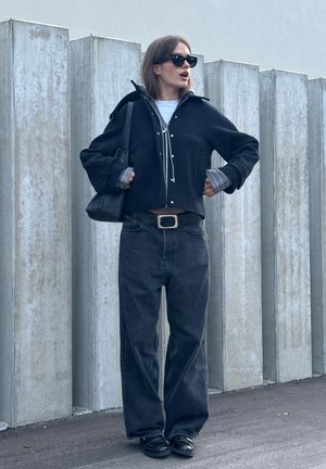 Chaqueta negra corta con botones plateados sobre una camisa blanca, combinada con jeans oscuros de pierna ancha y cintura alta, y zapatos negros sin cordones. Bolsa negra y gafas de sol.