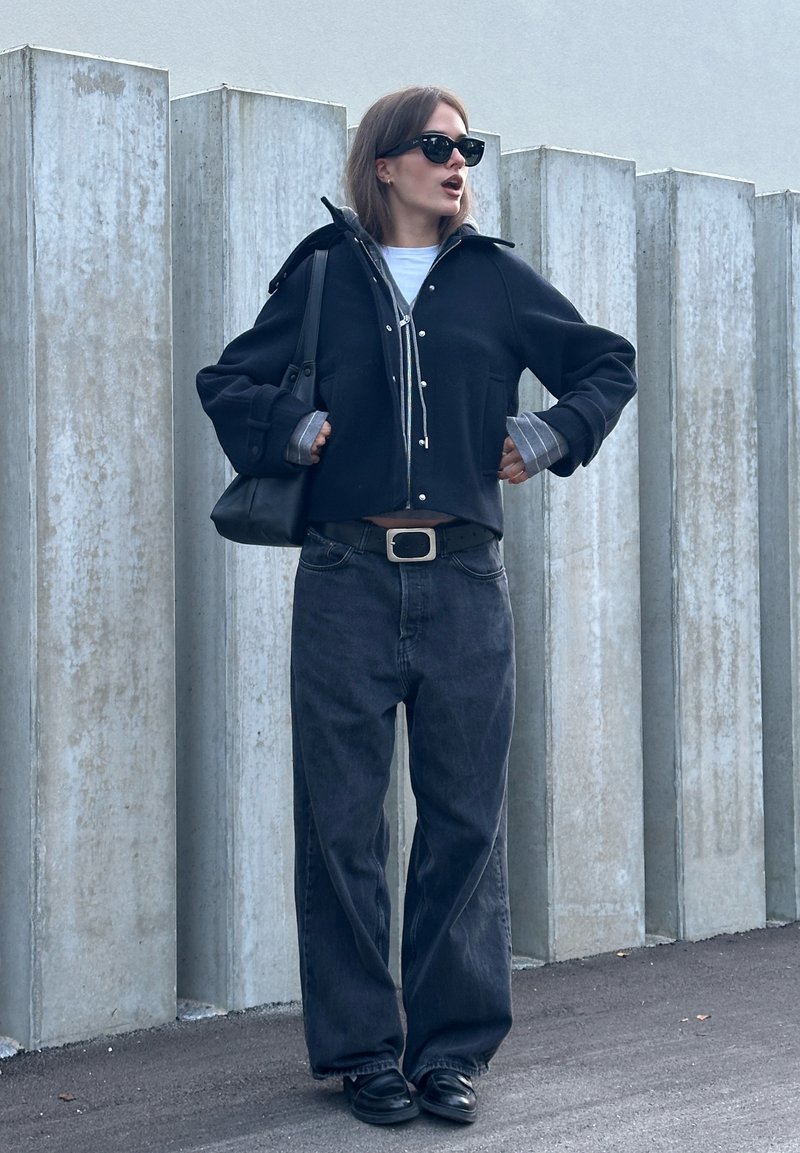 Corta giacca nera con bottoni argentati sopra una camicia bianca, abbinata a jeans scuri a vita alta e gamba larga, insieme a scarpe nere slip-on. Borsa nera e occhiali da sole.