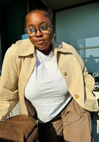 Μπεζ υπερμεγέθης ζακέτα με κουμπιά πάνω από ένα λευκό μπλουζάκι, συνδυασμένη με καφέ παντελόνι. Παχύ διάφανο γυαλιά και υφασμάτινη υφή.