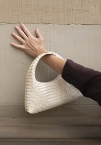 Hånd som hviler på beige steinvegg, iført mørkermede, holder en off-white vevd håndveske med rund håndtak over håndleddet.