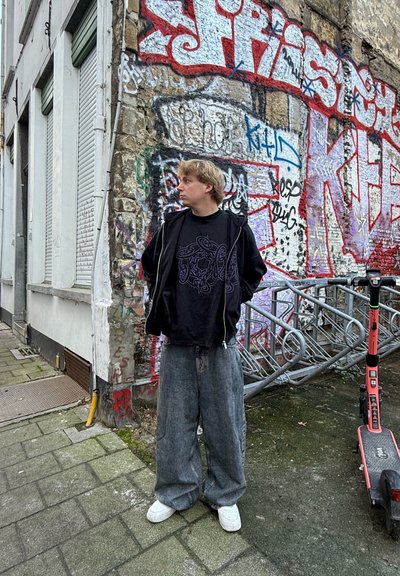 Sudadera negra con cremallera sobre una camiseta gráfica, combinada con jeans amplios grises y zapatillas deportivas blancas, apoyada contra una pared cubierta de graffiti.