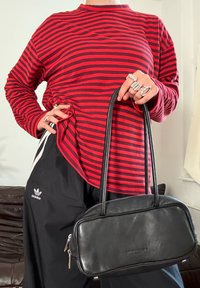 Chemise à manches longues rayée rouge et noire associée à un pantalon de sport noir. Un petit sac à main en cuir noir avec double poignée est tenu dans une main.