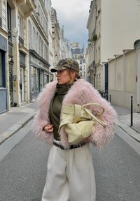 Femme en veste rose duveteuse, col roulé vert olive, casquette camouflage, pantalon blanc, tenant un sac à main beige, debout dans une rue étroite européenne.