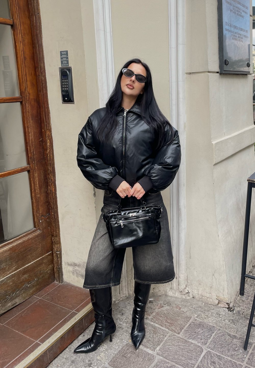 Giubbotto bomber in pelle nera con maniche gonfie, abbinato a pantaloni di jeans scuri al ginocchio e stivaletti neri lucidi. Accessorio: piccola borsa nera.