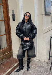 Giubbotto bomber in pelle nera con maniche gonfie, abbinato a pantaloni di jeans scuri al ginocchio e stivaletti neri lucidi. Accessorio: piccola borsa nera.