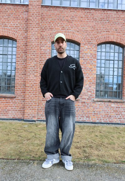 Hombre con cárdigan negro, vaqueros anchos descoloridos, zapatillas blancas y gorra verde, de pie sobre césped frente a un edificio de ladrillo rojo con ventanas arqueadas.