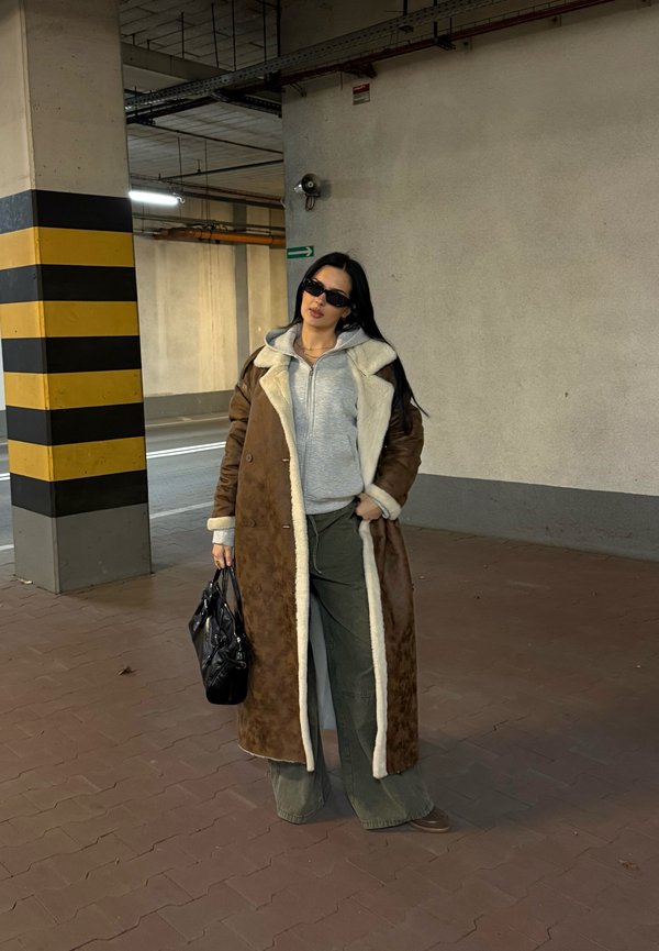 Cappotto di shearling marrone con rivestimento bianco, felpa grigia con zip, pantaloni verdi a gamba larga e borsa nera; indossando occhiali da sole scuri. Ambientazione in un parcheggio.