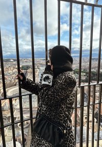 Personne en manteau à imprimé léopard et capuche noire tenant des barreaux métalliques, surplombant un paysage urbain sous un ciel nuageux.