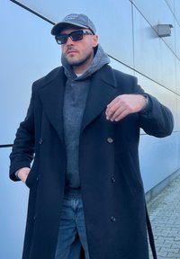 Μαύρο διπλό περαστό παλτό πάνω από γκρι φούτερ, συνδυασμένο με σκούρα τζιν. Φοράει γκρι καπέλο και μαύρα γυαλιά ηλίου, με τα χέρια στις τσέπες.