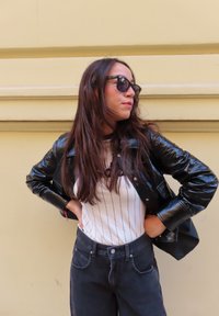 Černá lakovaná kožená bunda, bílá pruhovaná tričko, vysoké černé džíny, dlouhé hnědé vlasy a černé sluneční brýle na světlém žlutém pozadí.