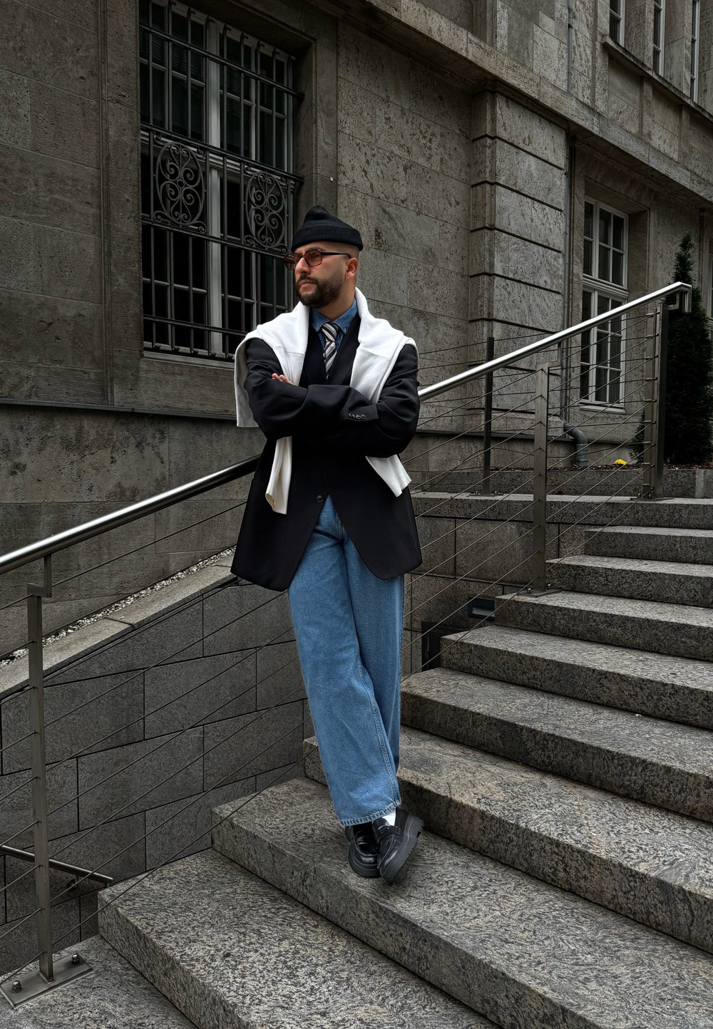 Άτομο φορώντας μαύρο σακάκι, ανοιχτό μπλε παντελόνι wide-leg και λευκή μπλούζα ριγμένη στους ώμους, στέκεται σε πέτρινα σκαλιά με μια κουπαστή.